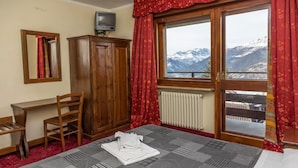 Une chambre d’hôtel comprenant un lit, une armoire en bois, un bureau, une chaise, une fenêtre donnant sur la montagne et un radiateur.