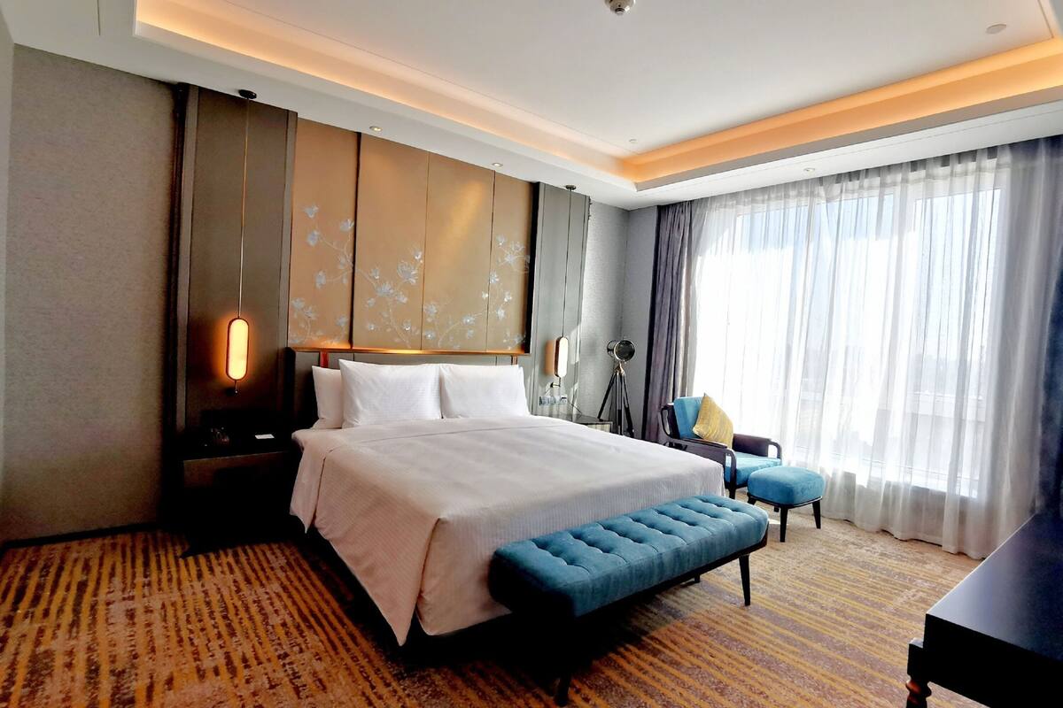A hotel room with a large bed, a bench, a desk, and a view of the city through the window.