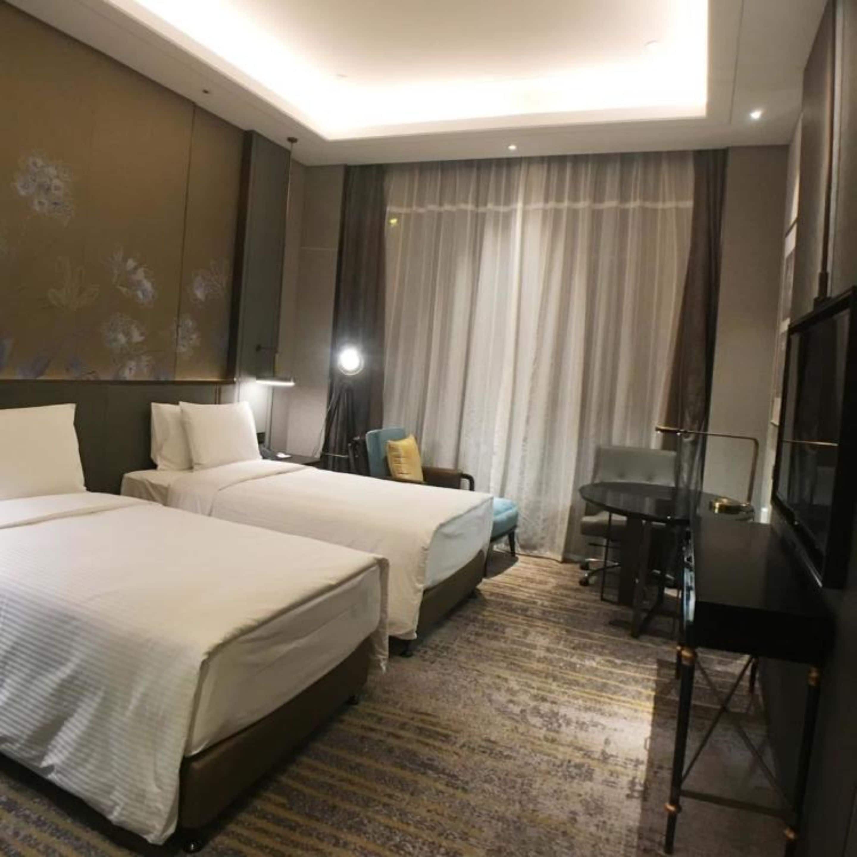 Habitación de hotel con dos camas, un escritorio, una silla, un televisor y un ventanal grande con cortinas.