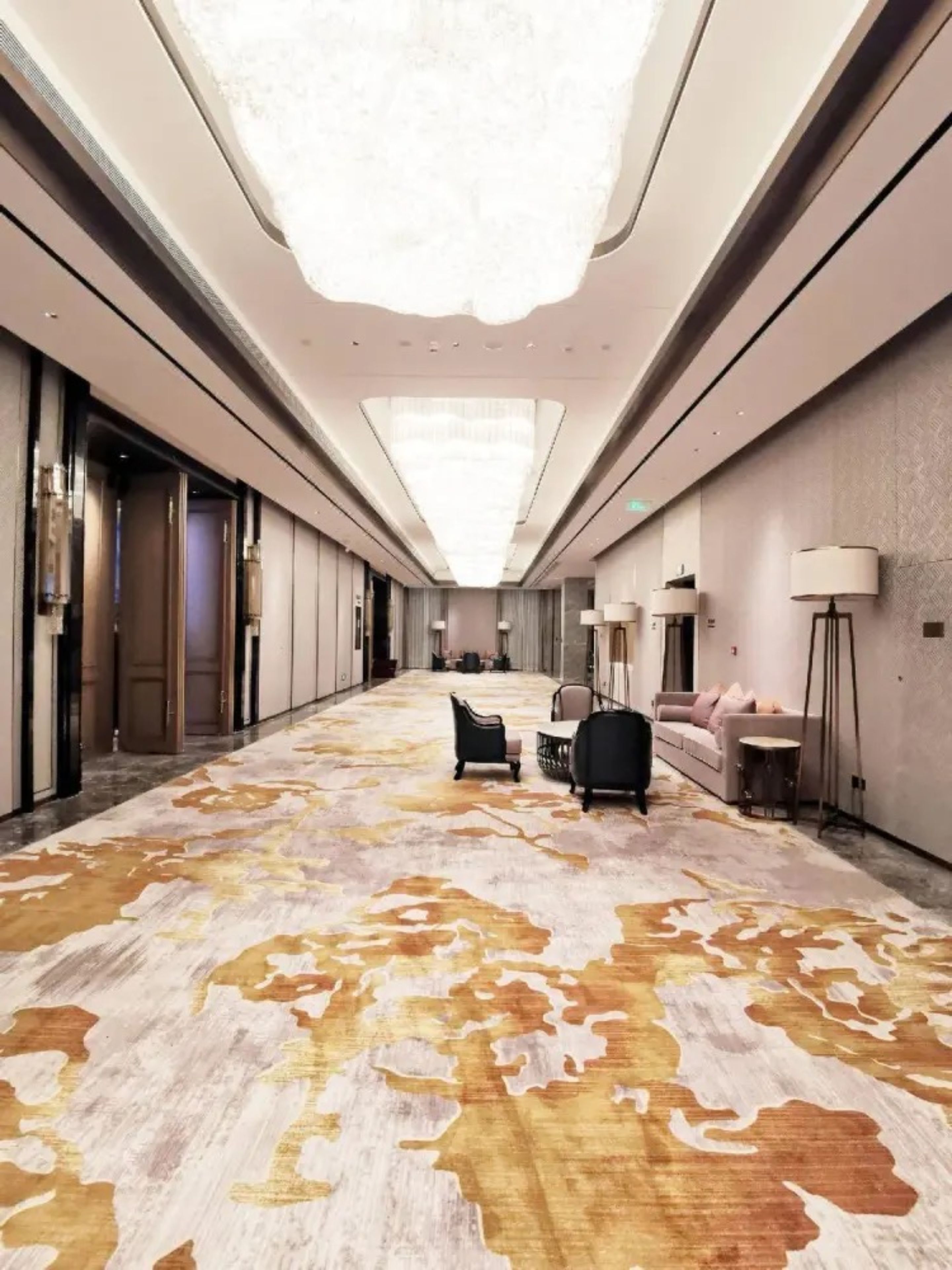 Un pasillo de hotel con alfombra estampada, rincones de descanso e iluminación moderna.