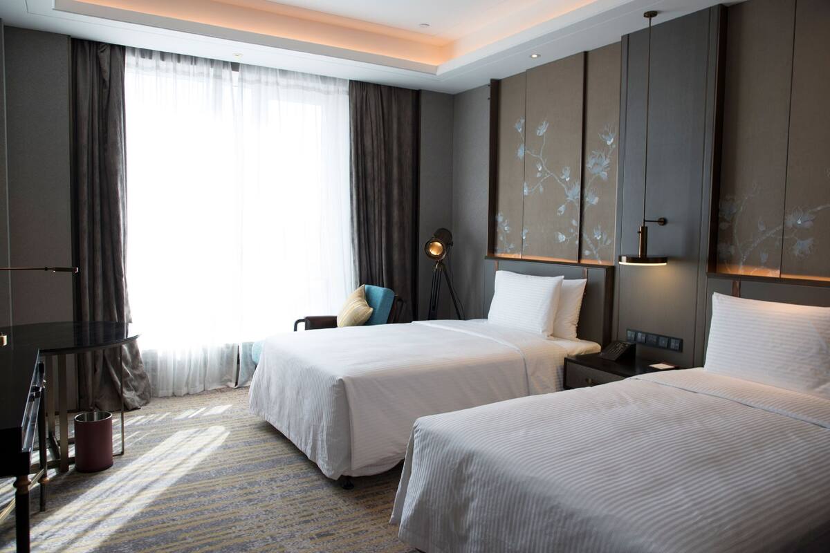A hotel room with two beds, a desk, a chair, a lamp, and a view of the outdoors.