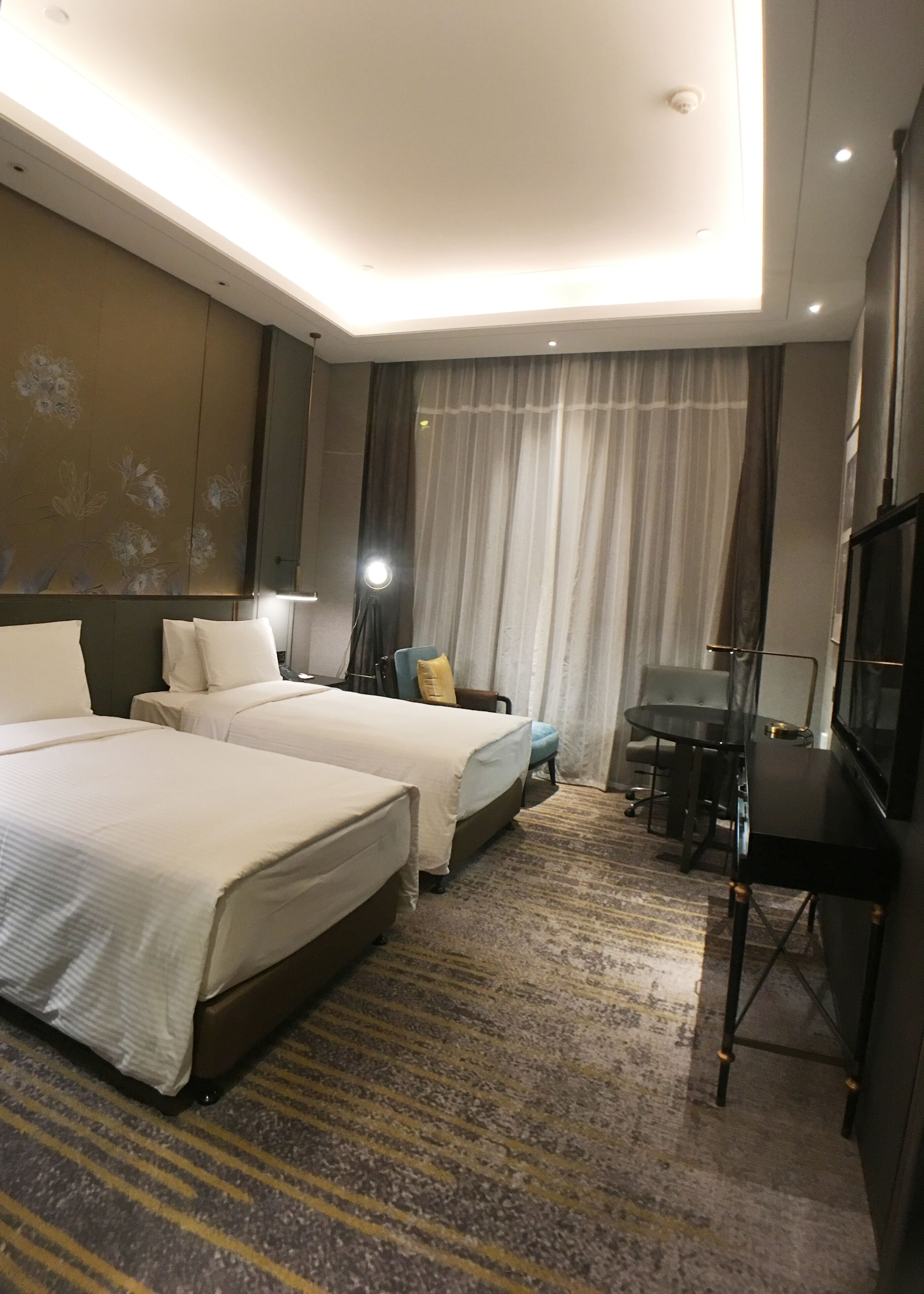 Habitación de hotel con dos camas, un escritorio, una silla, un televisor y un amplio ventanal con cortinas.