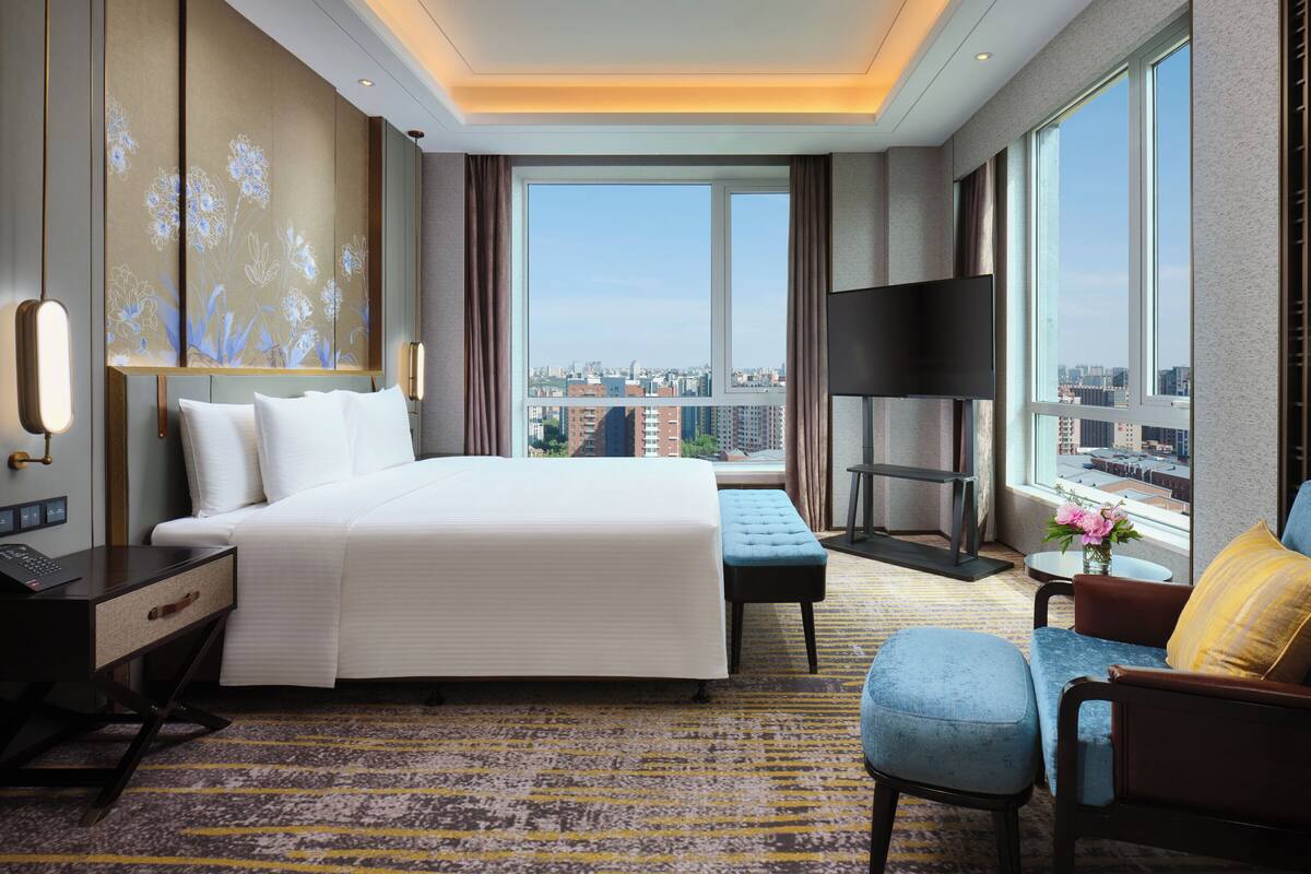 A modern hotel room with a large bed, a desk, a chair, a TV, and a view of the city.