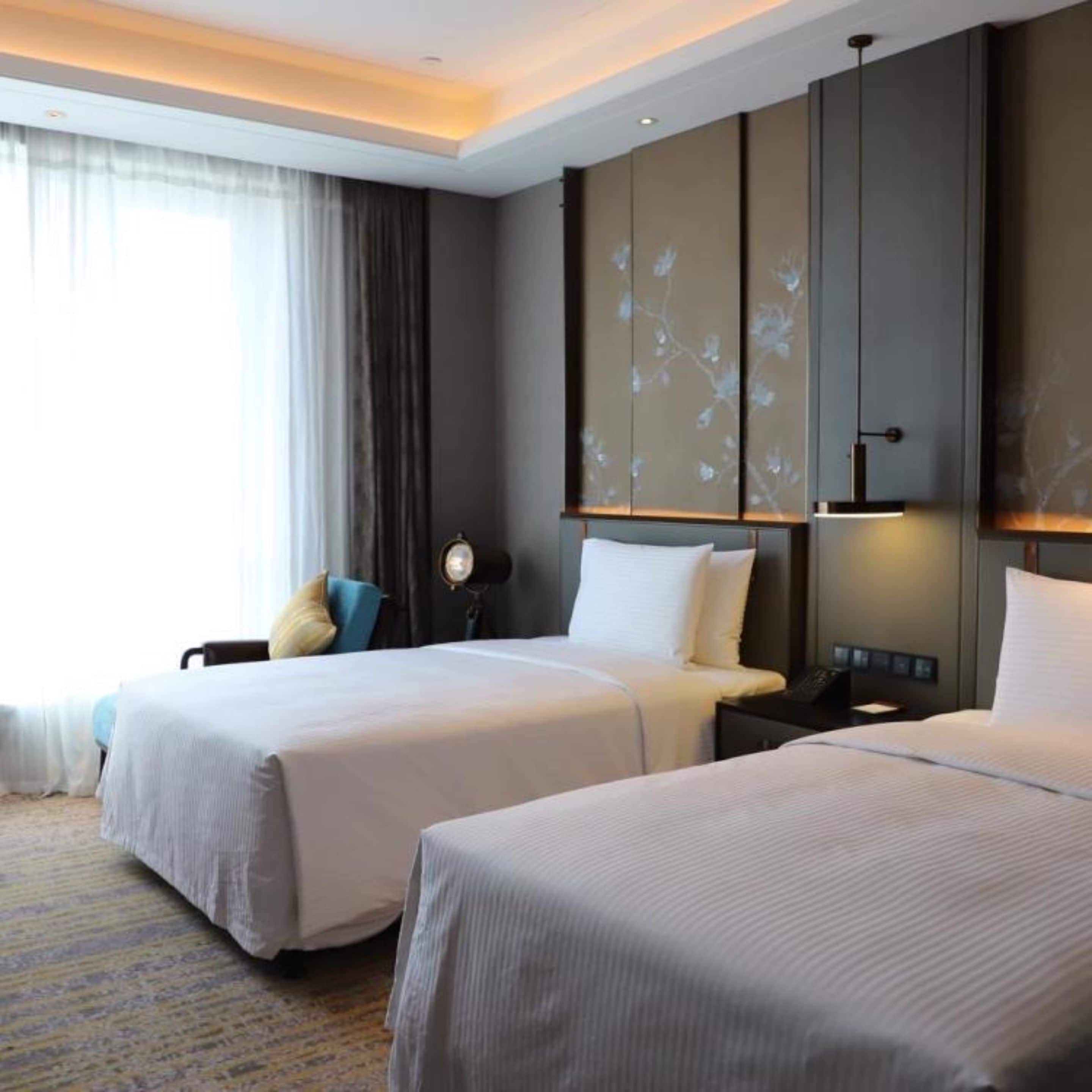 Habitación de hotel con dos camas, un escritorio, una silla y un ventanal con cortinas.