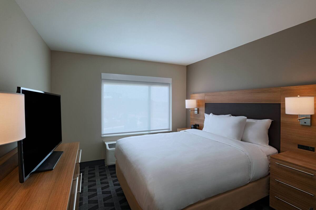 Habitación de hotel con una cama grande, un televisor de pantalla plana, una ventana y una mesita de noche con lámpara.