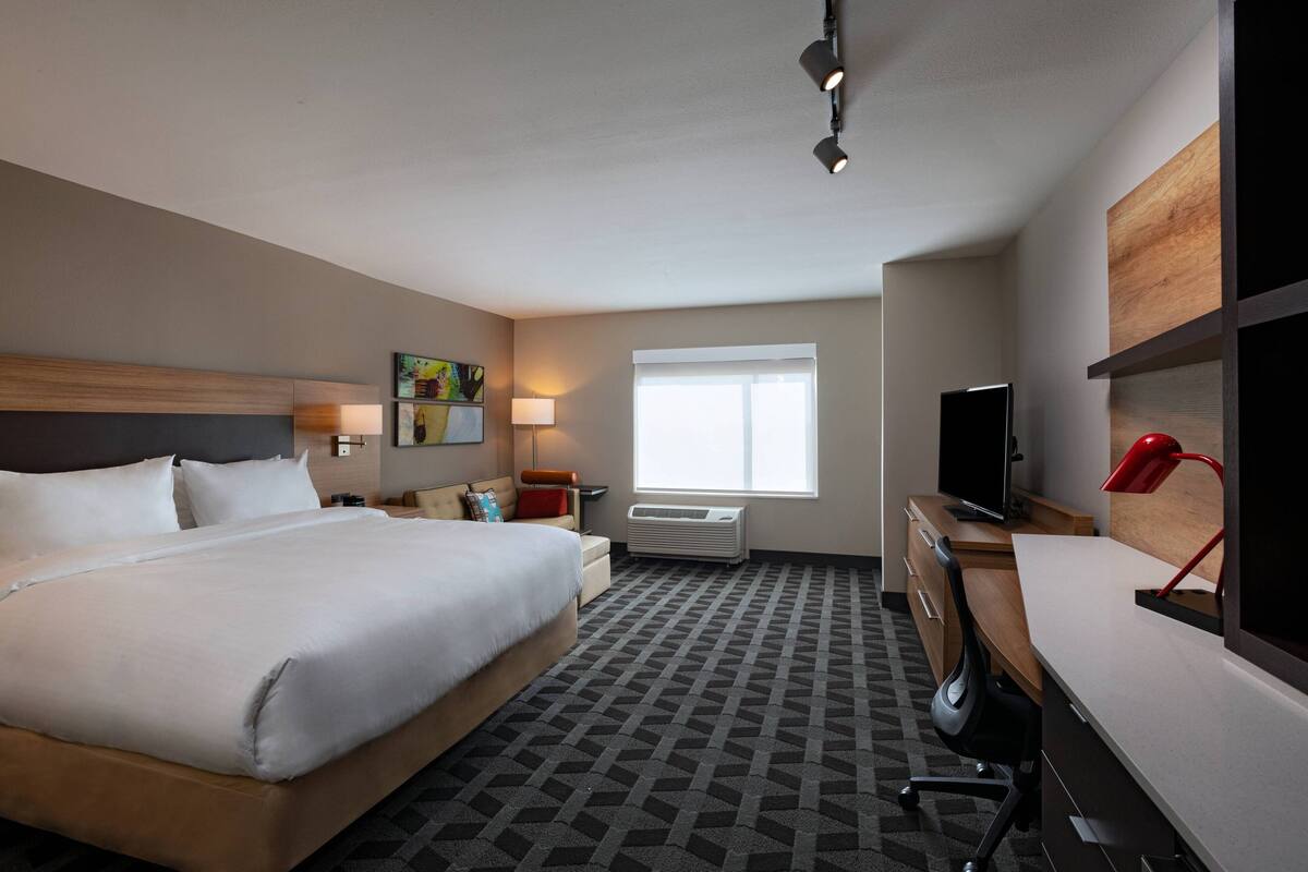 Habitación de hotel con una cama grande, un escritorio con computadora, televisión, ventana y estante montado en la pared.