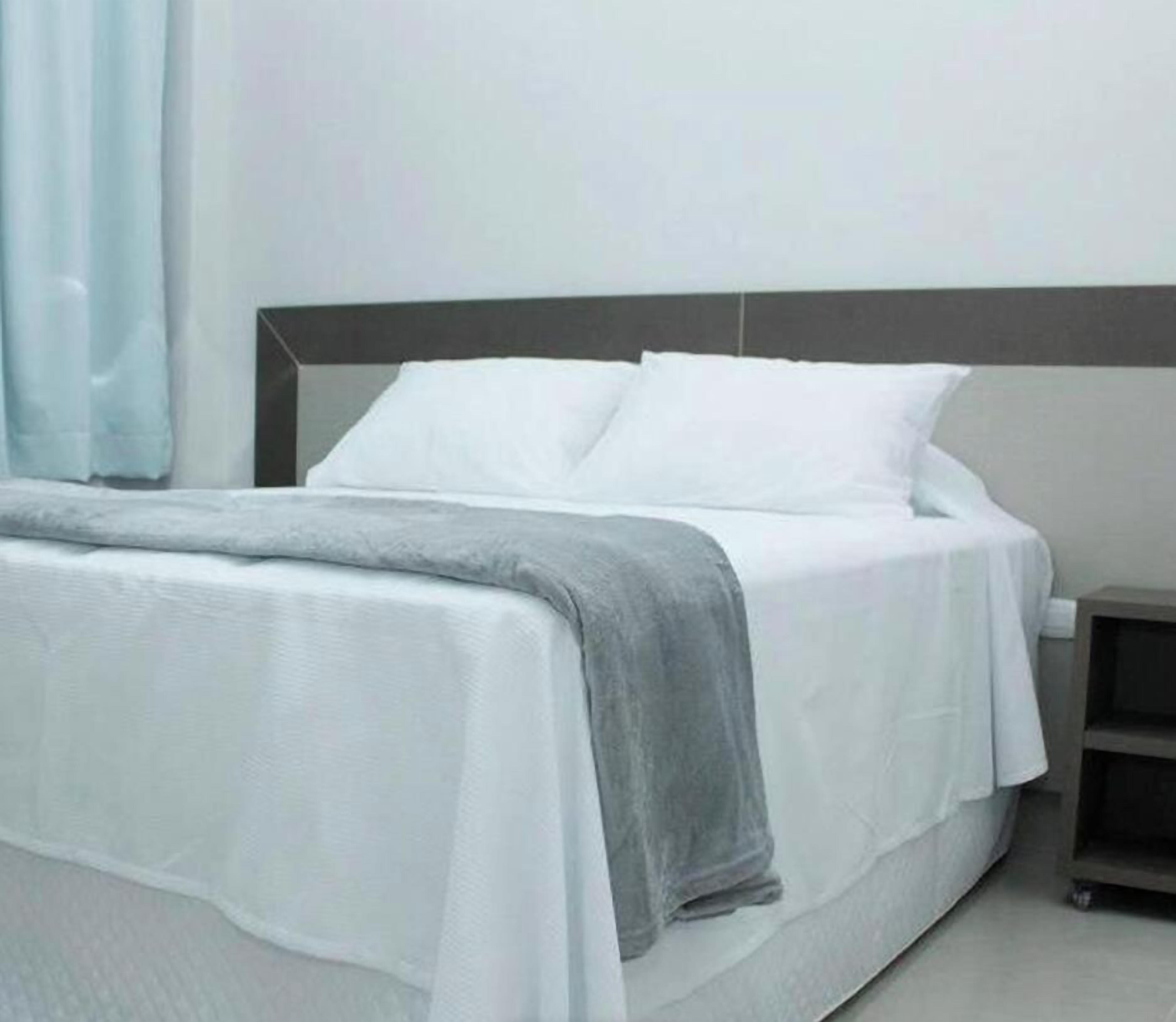 Un lit avec du linge de lit blanc et un plaid gris, une table de chevet et une commode.