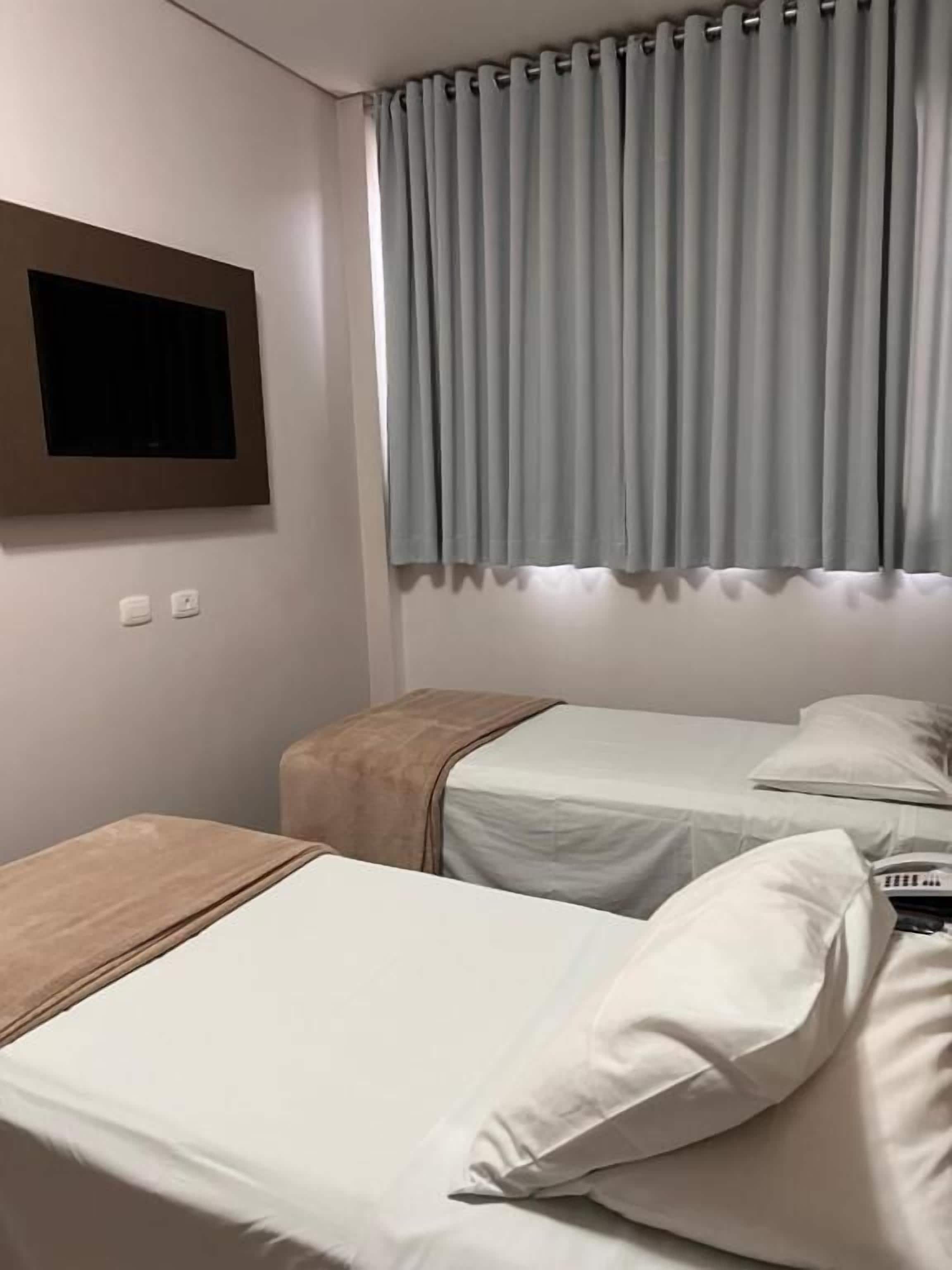 A hotel room with two beds, a flat-screen TV mounted on the wall, and a window with curtains.