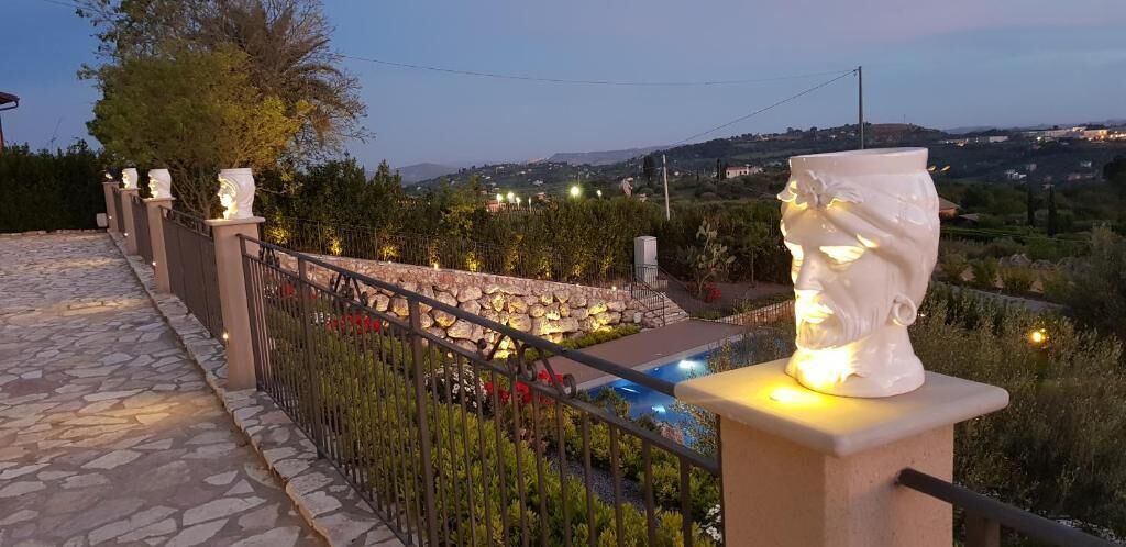 Un chemin de pierre mène à un espace piscine agrémenté d’une sculpture décorative représentant une tête et offrant une vue sur la campagne environnante.