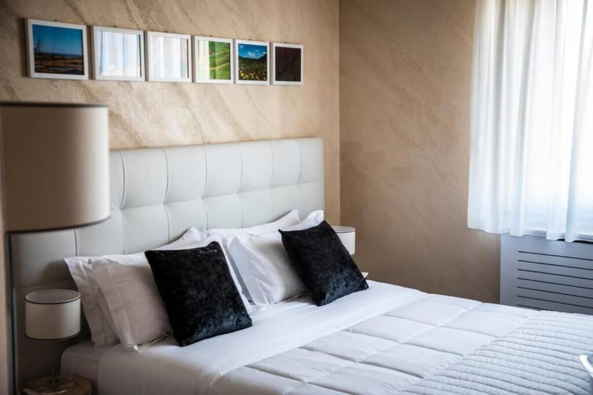 Habitació d'hotel amb un llit blanc, almoinades negres, una taula de nit amb una làmpada i una paret amb quadres.