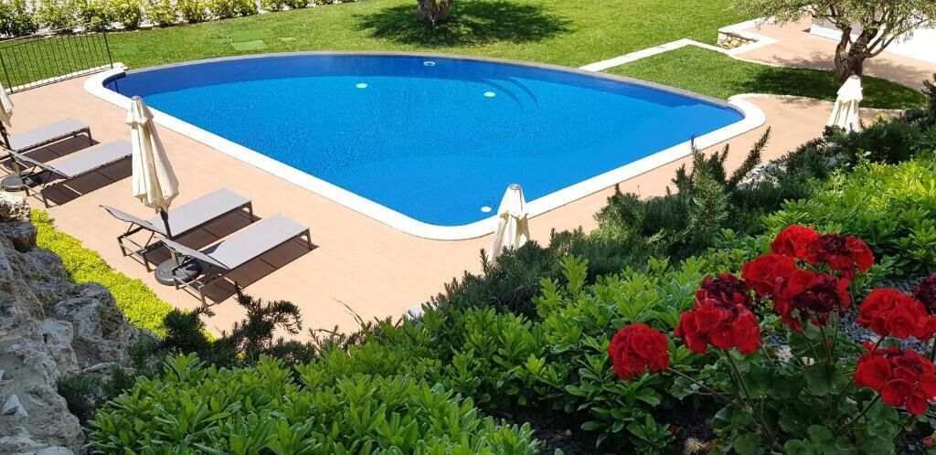 Una piscina rettangolare con interno blu, circondata da un solarium color beige. Lungo il bordo della piscina si trovano sedie a sdraio e ombrelloni.