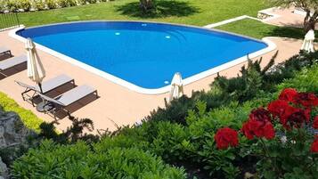Una piscina rettangolare con interno blu, circondata da un solarium color beige. Lungo il bordo della piscina si trovano sedie a sdraio e ombrelloni.