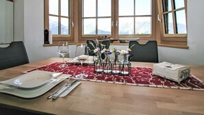 Comedor con una mesa de madera dispuesta para cuatro, ventanas amplias con vistas a las montañas y un jarrón con flores sobre la mesa.