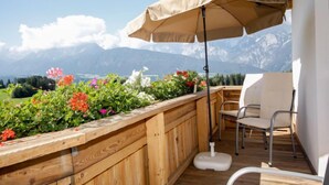 Una terraza de madera con un banco y una silla, una mesa con sombrilla blanca, y un cantero con diversas flores.