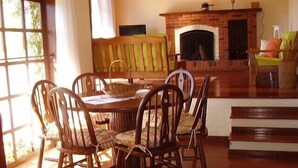 A dining area with a wooden table and chairs, a brick fireplace, and a view of the outdoors through a large window.