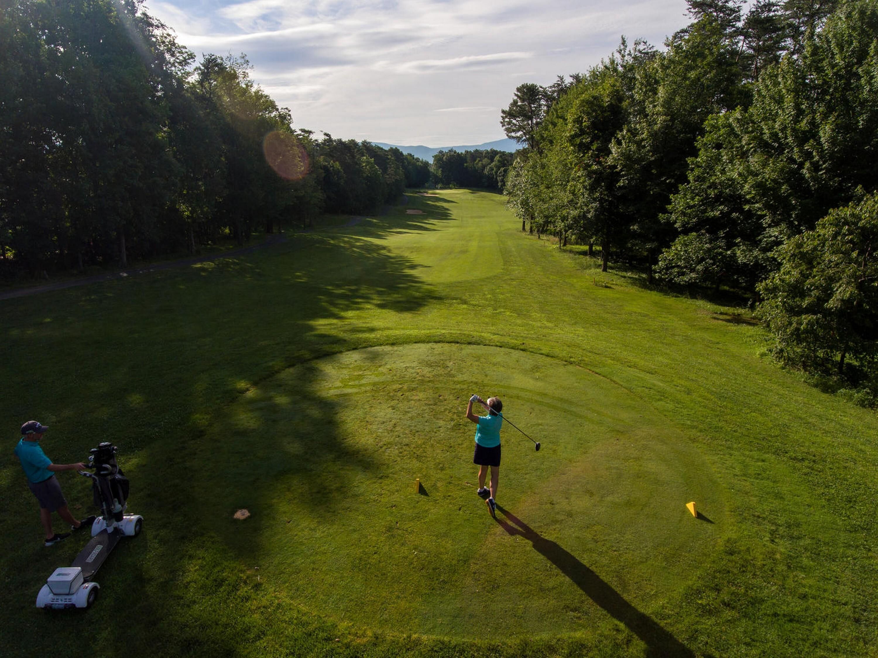 Campo de golf con un sendero despejado, árboles y una persona con camisa azul preparándose para golpear una pelota.