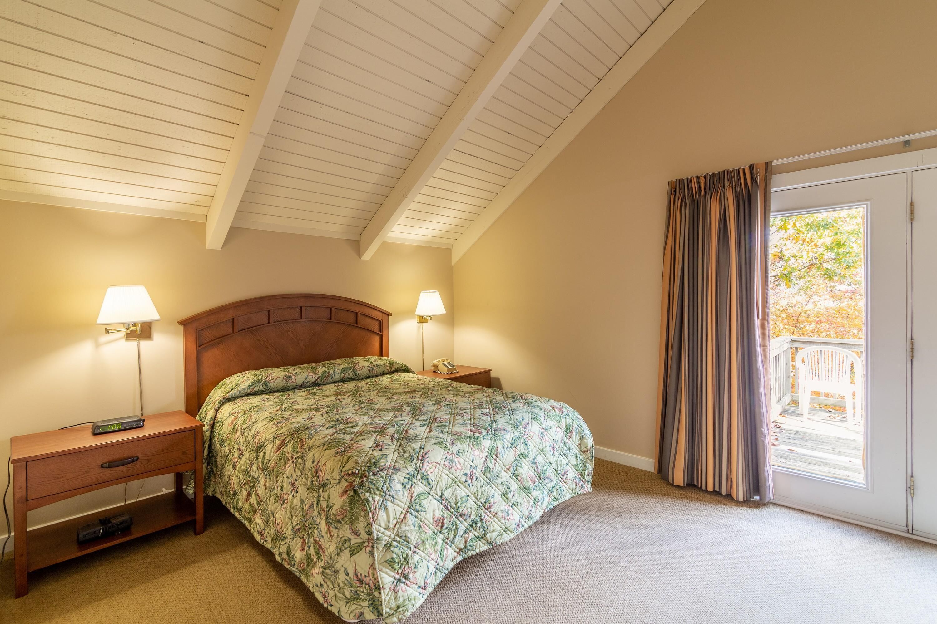 A bedroom with a wooden headboard, a bed with a floral comforter, two bedside lamps, a nightstand with a drawer, and a sliding glass door leading to an outdoor area.