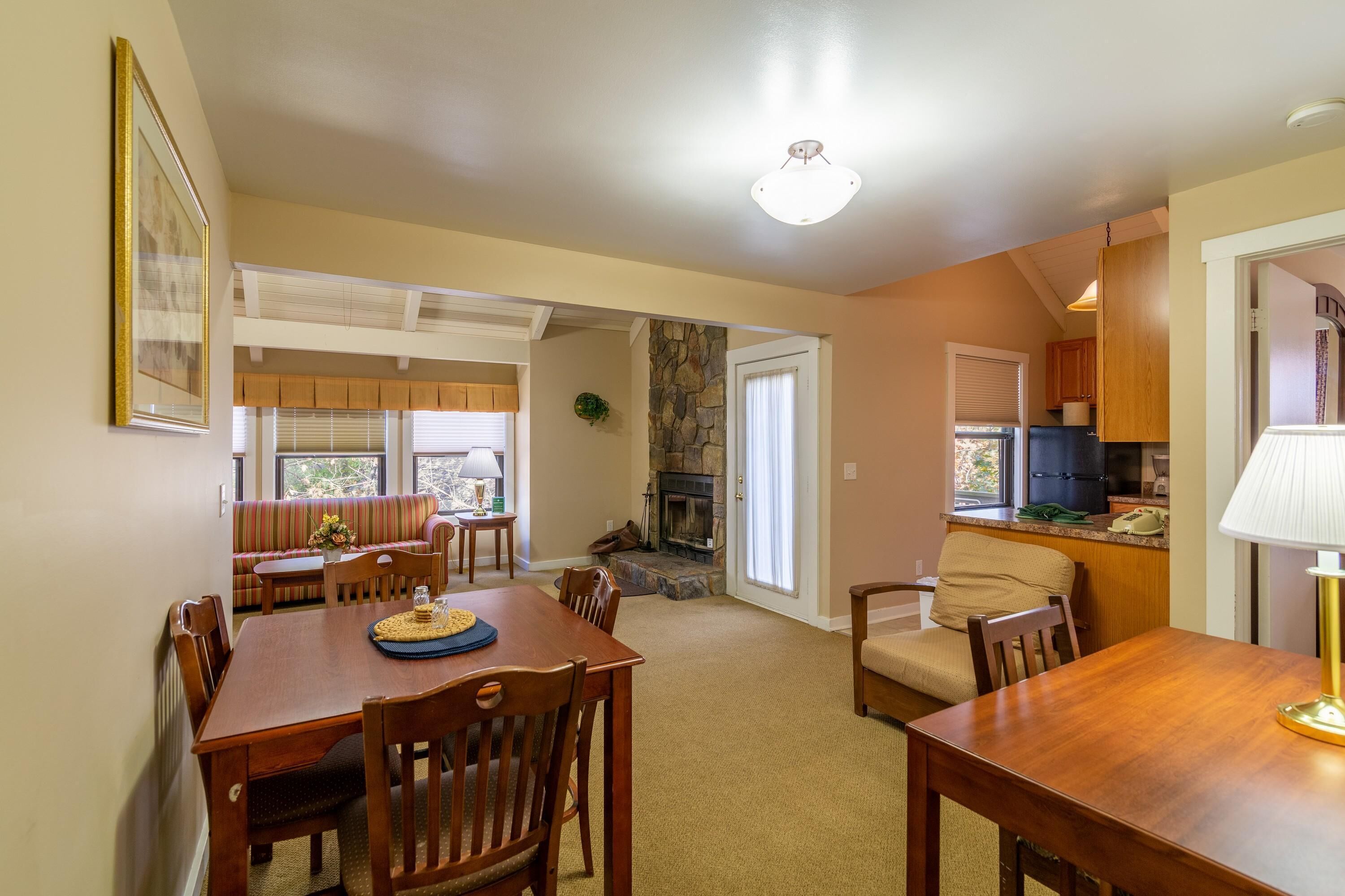 A dining area with a wooden table and chairs, a stone fireplace, and a kitchen in the background.