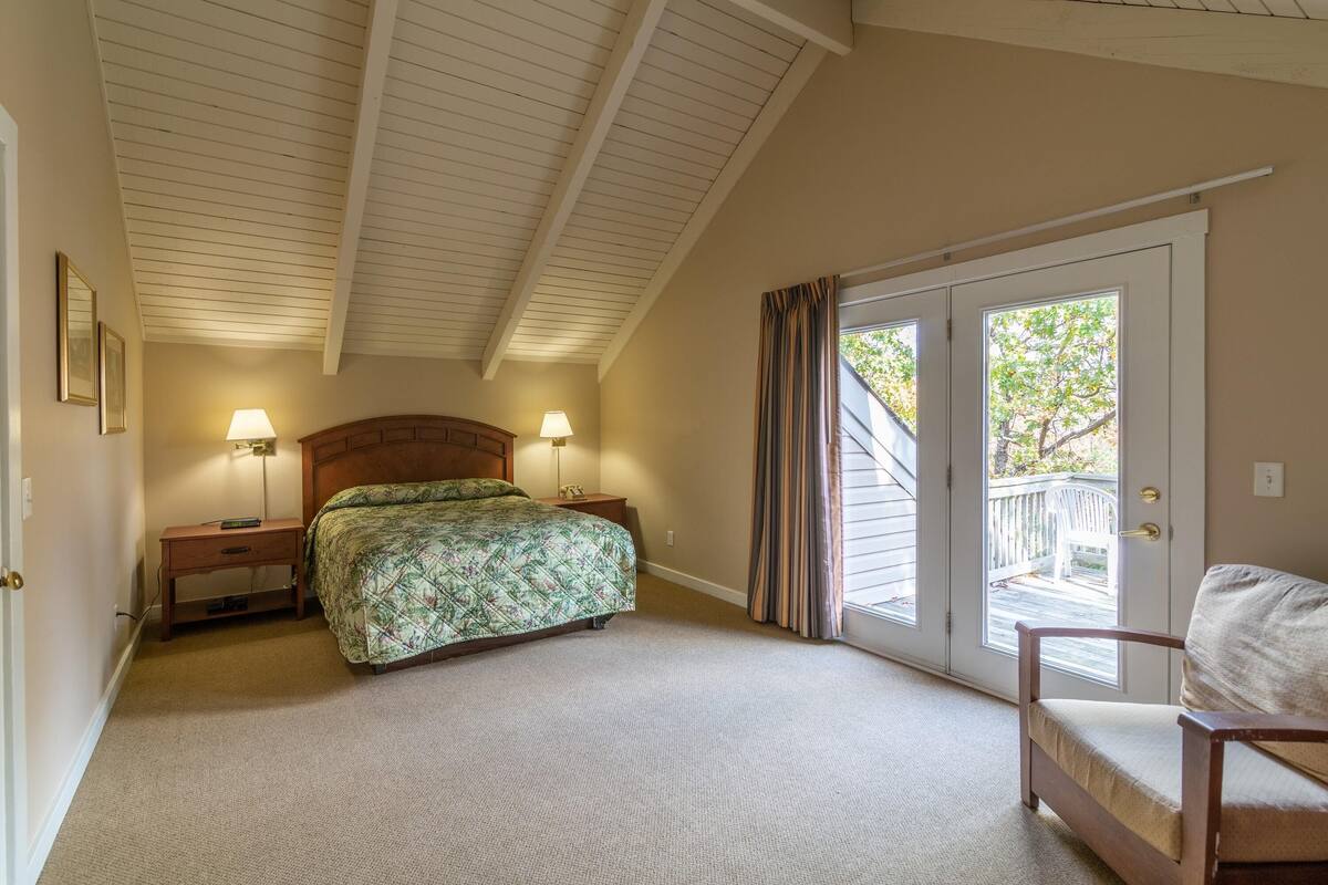 Une chambre à coucher comprenant un lit, deux lampes de chevet, une table de nuit en bois, une chaise et une porte-fenêtre coulissante donnant sur un balcon.