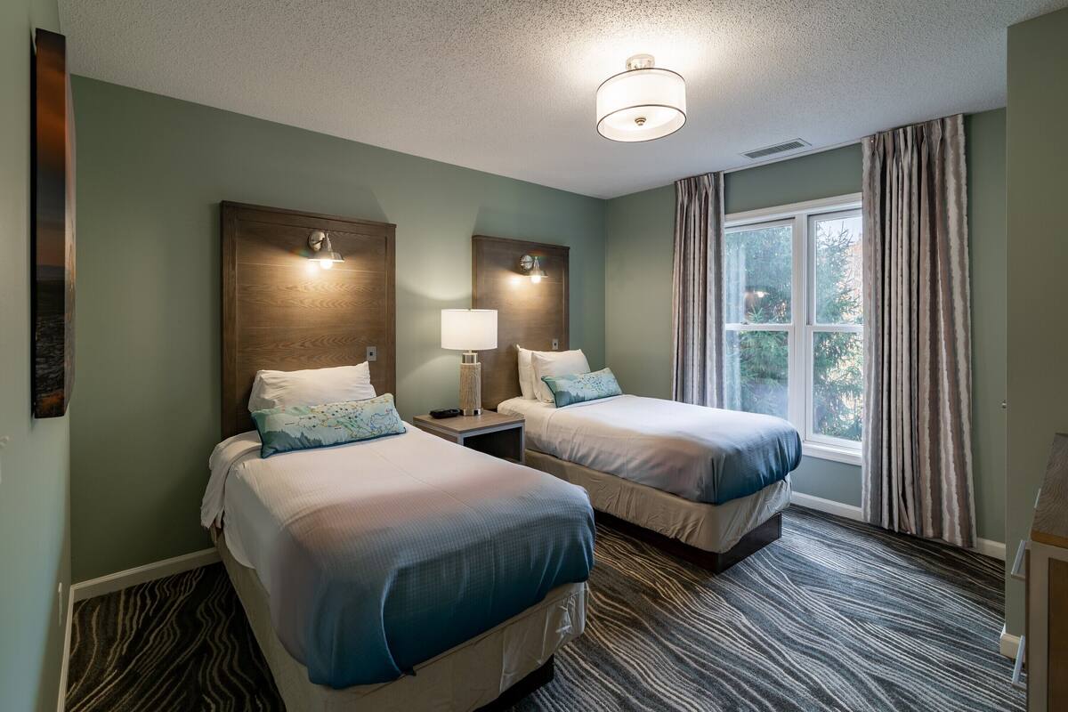 Habitación de hotel con dos camas, una ventana con cortinas, una lámpara en cada mesita de noche y una alfombra con textura.