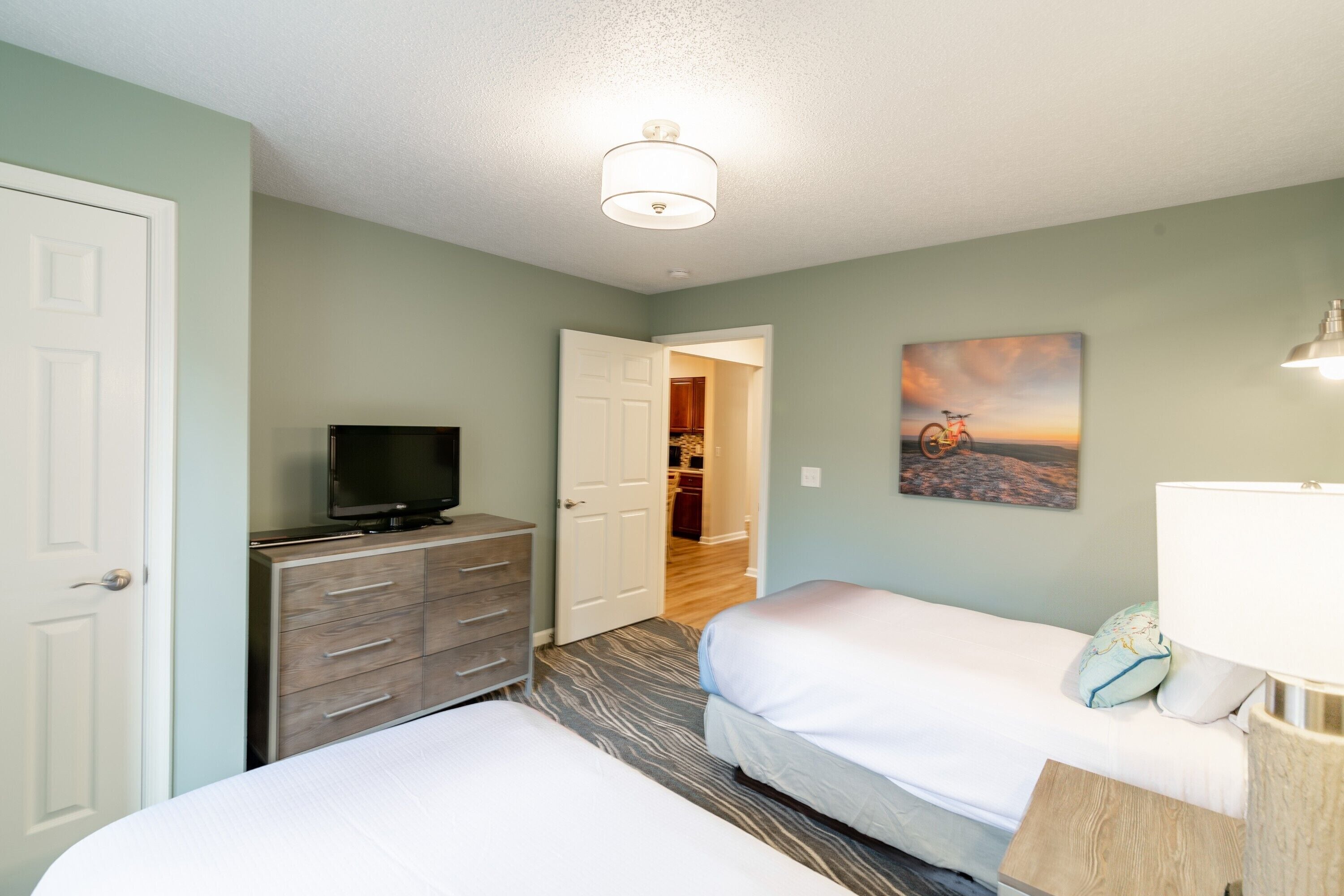 A hotel room with two beds, a television on a dresser, a painting on the wall, and a lamp on the side table.