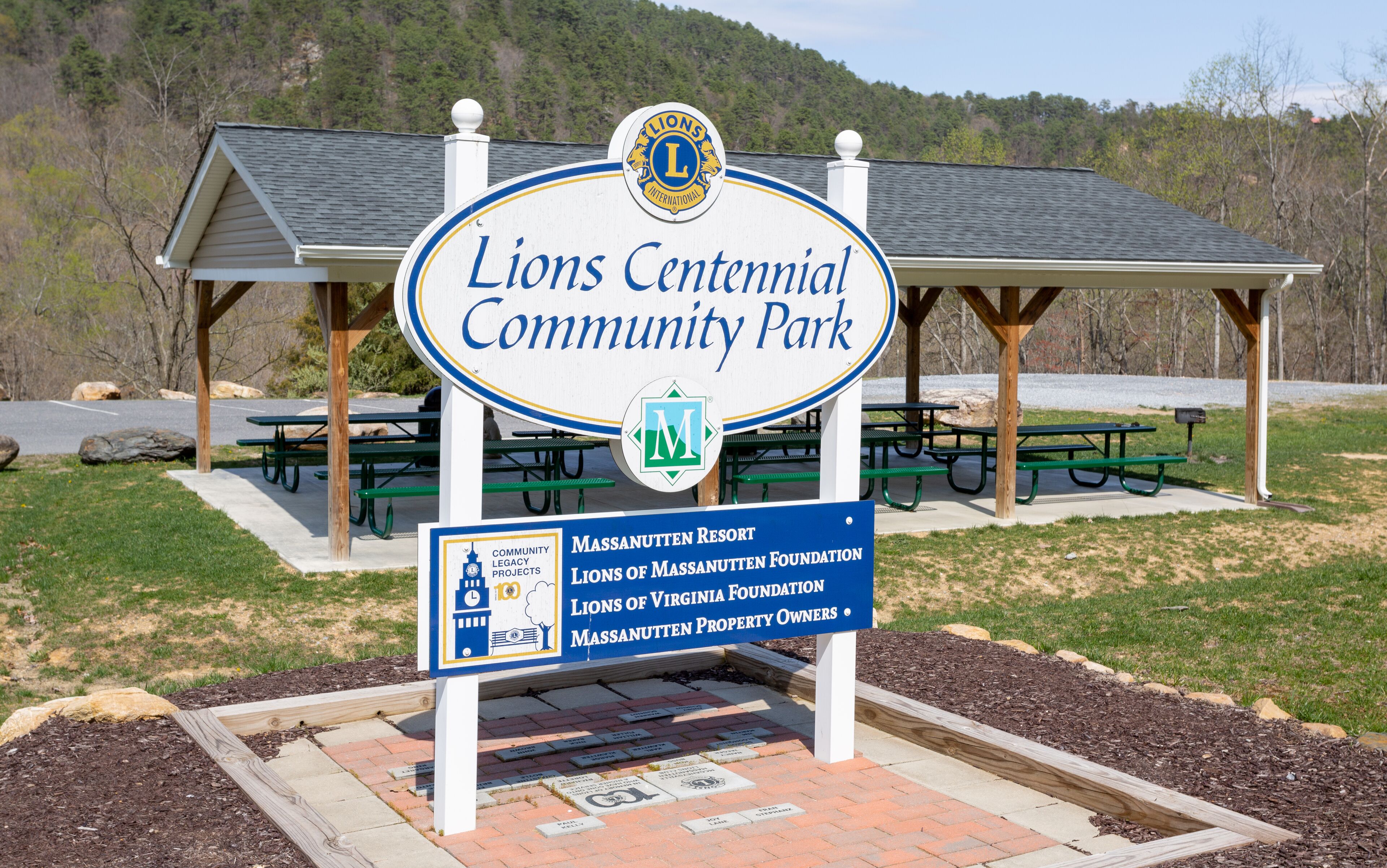 Letrero del Parque Comunitario Lions Centennial, con mesas de picnic y un refugio al fondo.
