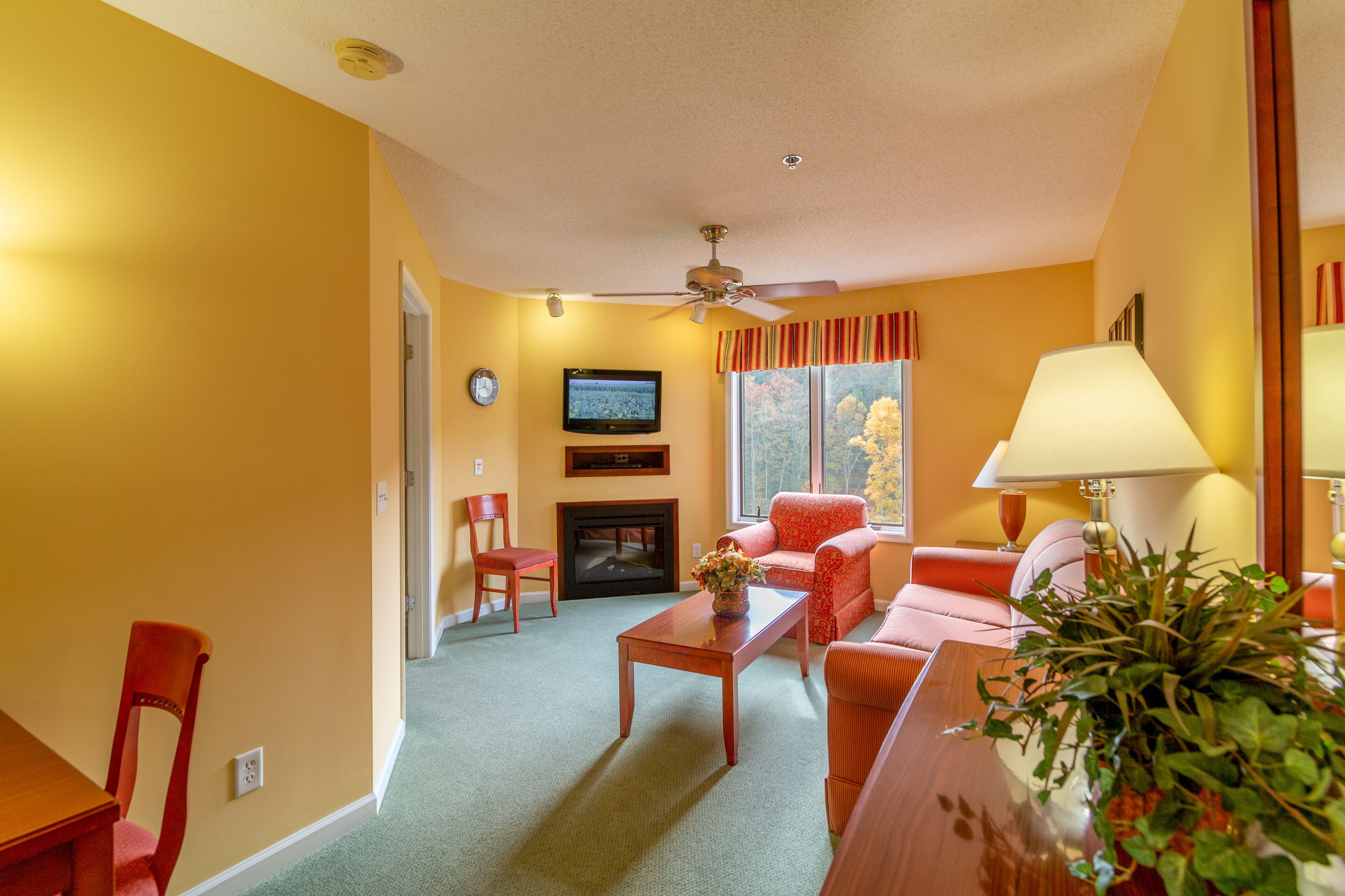 A living room with a fireplace, a television, a ceiling fan, and a window with curtains.