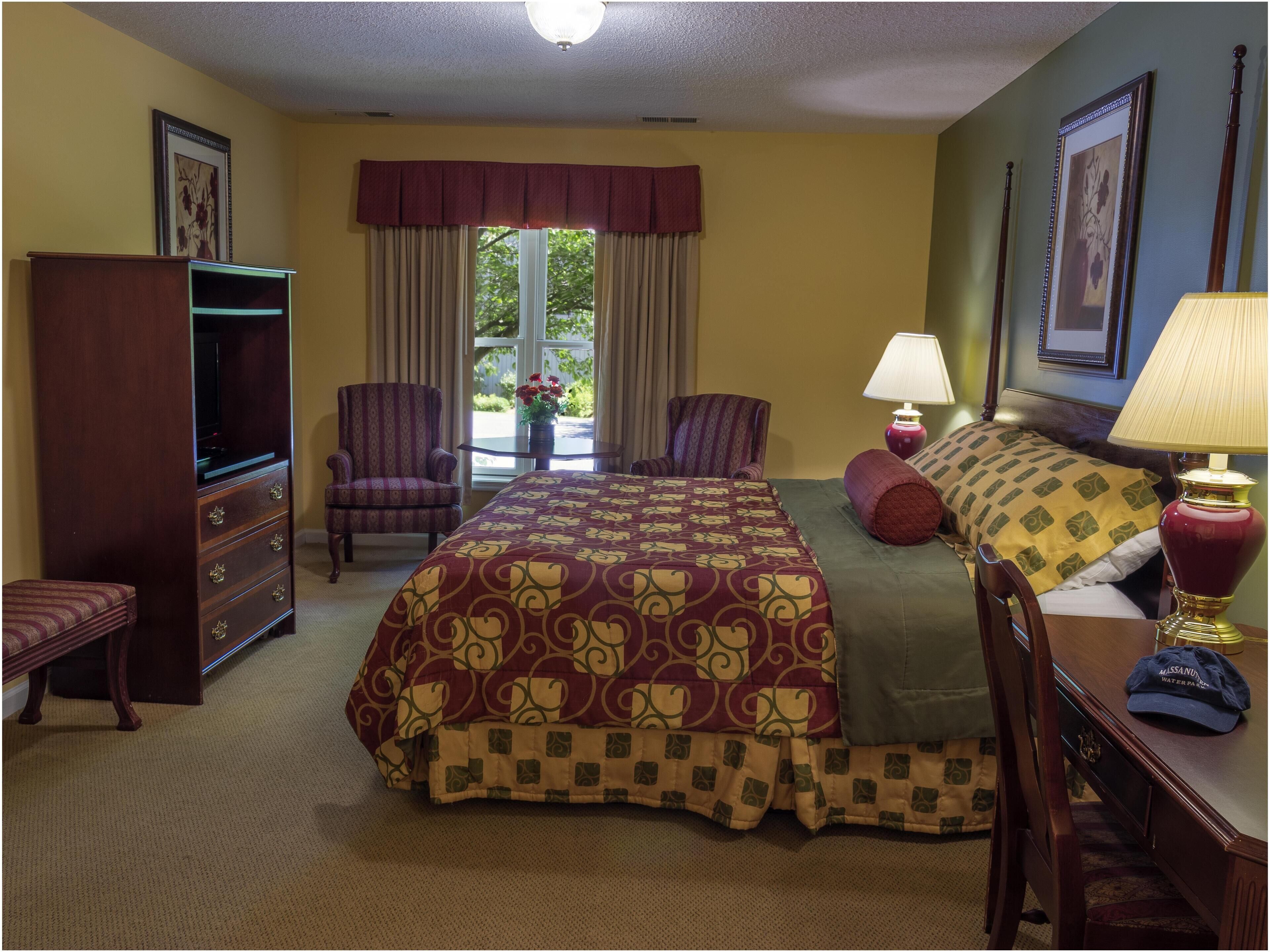 Une chambre d’hôtel avec un grand lit, deux fauteuils, une commode, une télévision et une fenêtre avec des rideaux.
