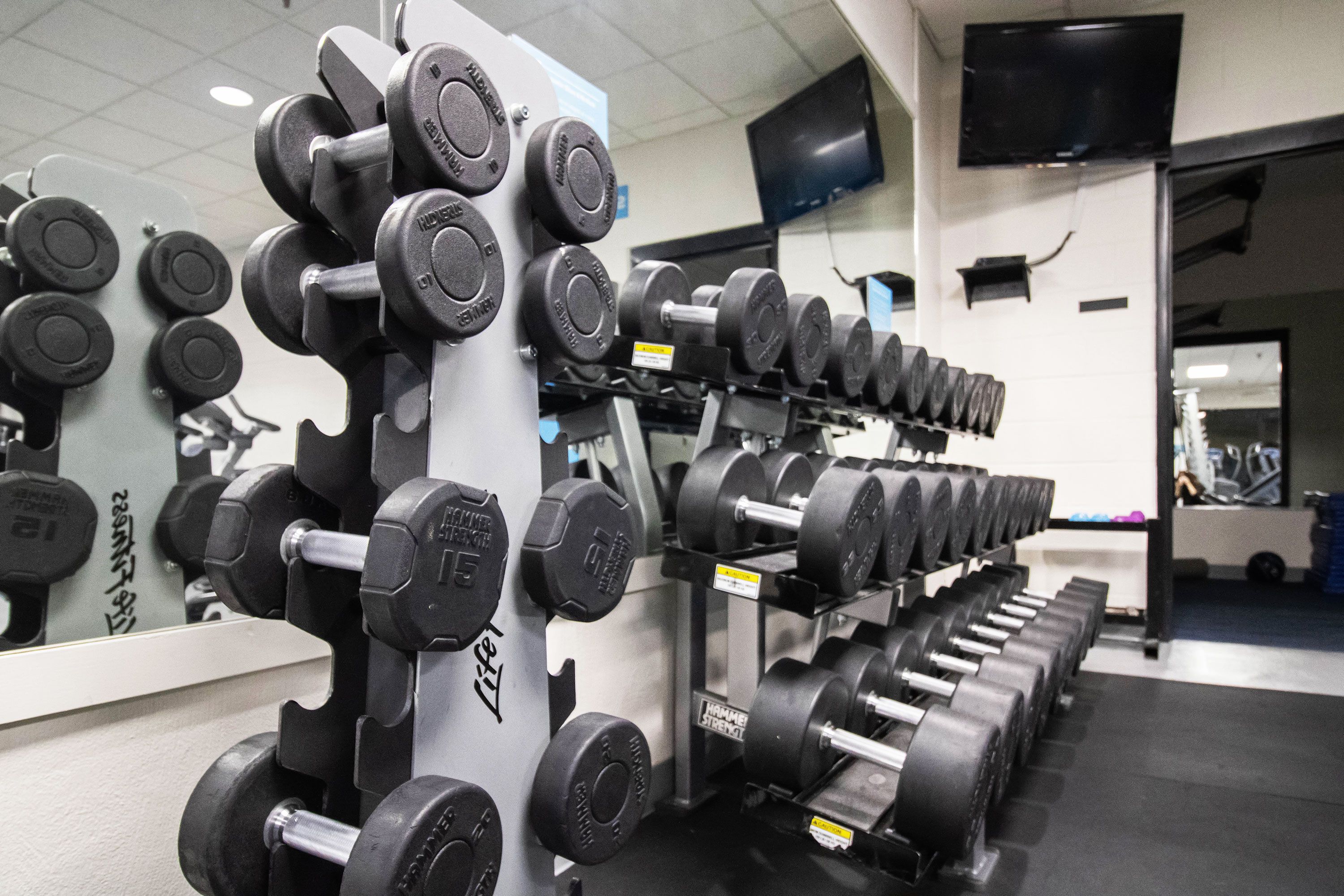 Une salle de sport avec des rangées d’haltères et de disques de poids, un téléviseur accroché au mur et un miroir qui reflète l’équipement.