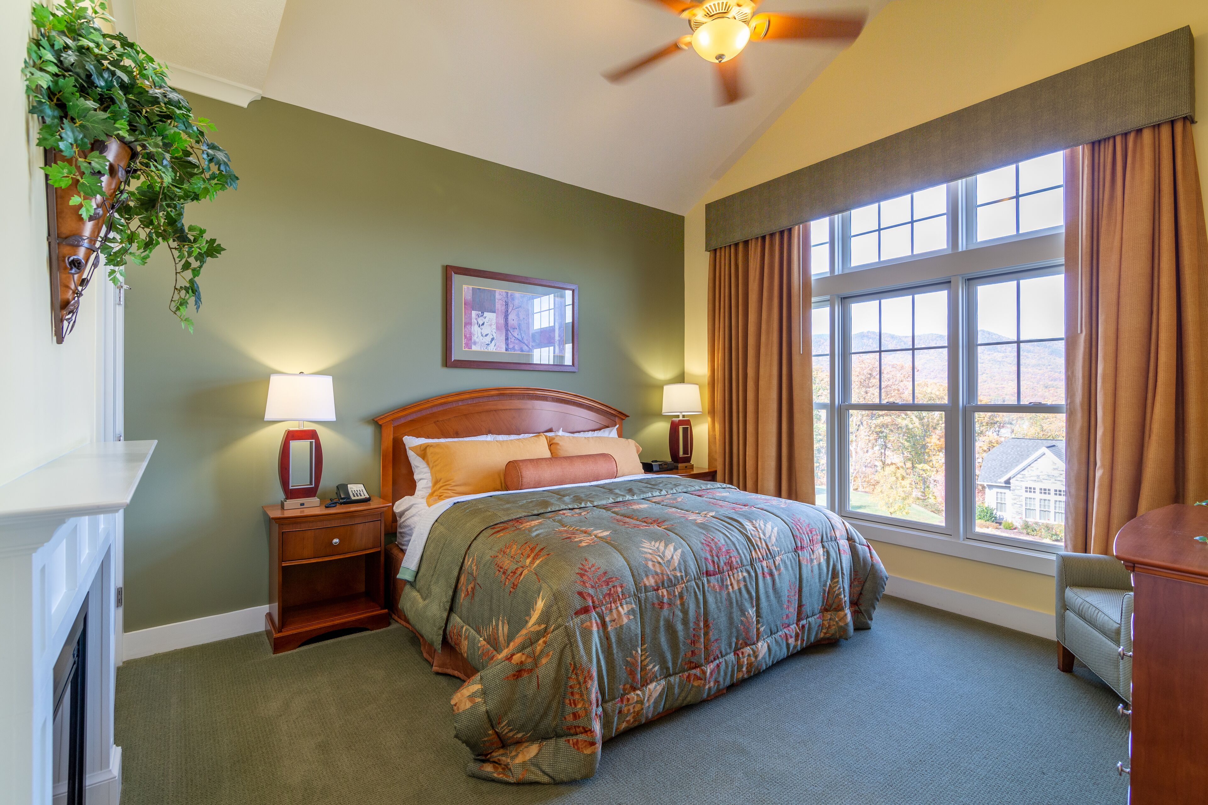 A bedroom with a large bed, two bedside tables with lamps, a ceiling fan, a window with curtains, and a wall-mounted plant.