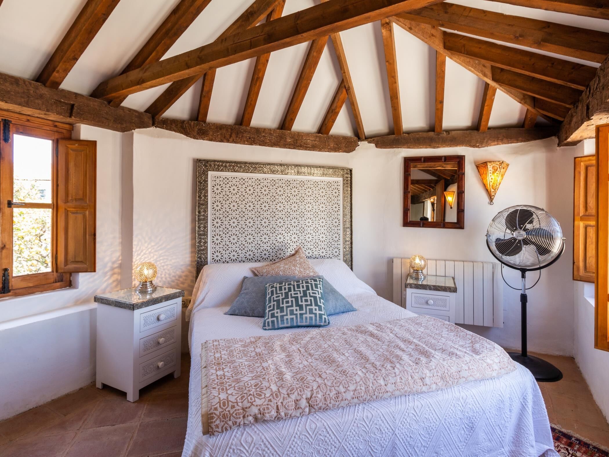 A bedroom with a large bed, wooden beams, a patterned headboard, bedside lamps, a floor fan, and a window with shutters.