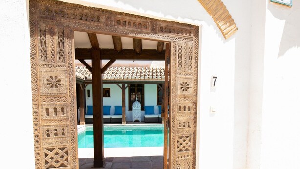 Ein Portal mit aufwendigen Schnitzereien führt zu einem Poolbereich mit einer gefliesten Dachterrasse und einem weißen Gebäude im Hintergrund.