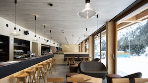 A modern bar with wooden stools and tables, large windows with a snowy view, and pendant lighting. - Campra Alpine Lodge & Spa (Blenio)