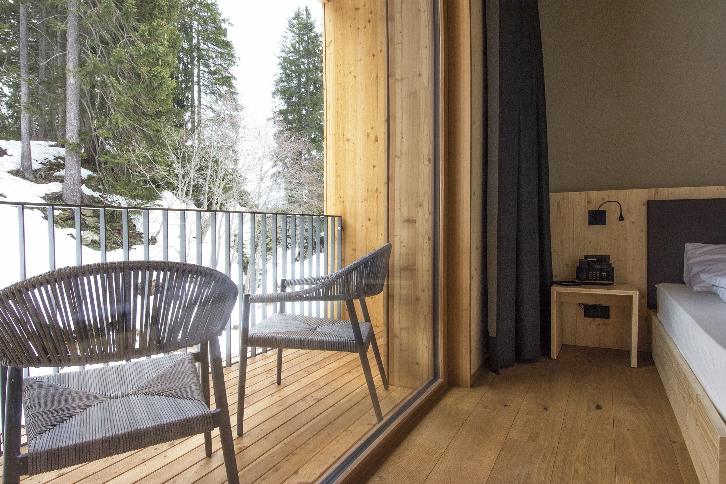 A wooden deck with two wicker chairs, a wooden nightstand with a phone, and a bed with white linens.