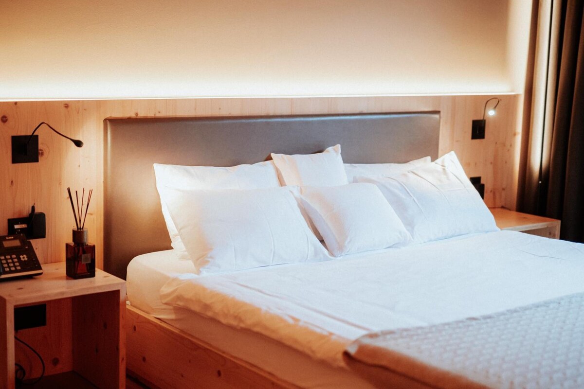 Una cama bien hecha con sábanas blancas, cabecera de madera y mesita de noche con una lámpara y un teléfono.