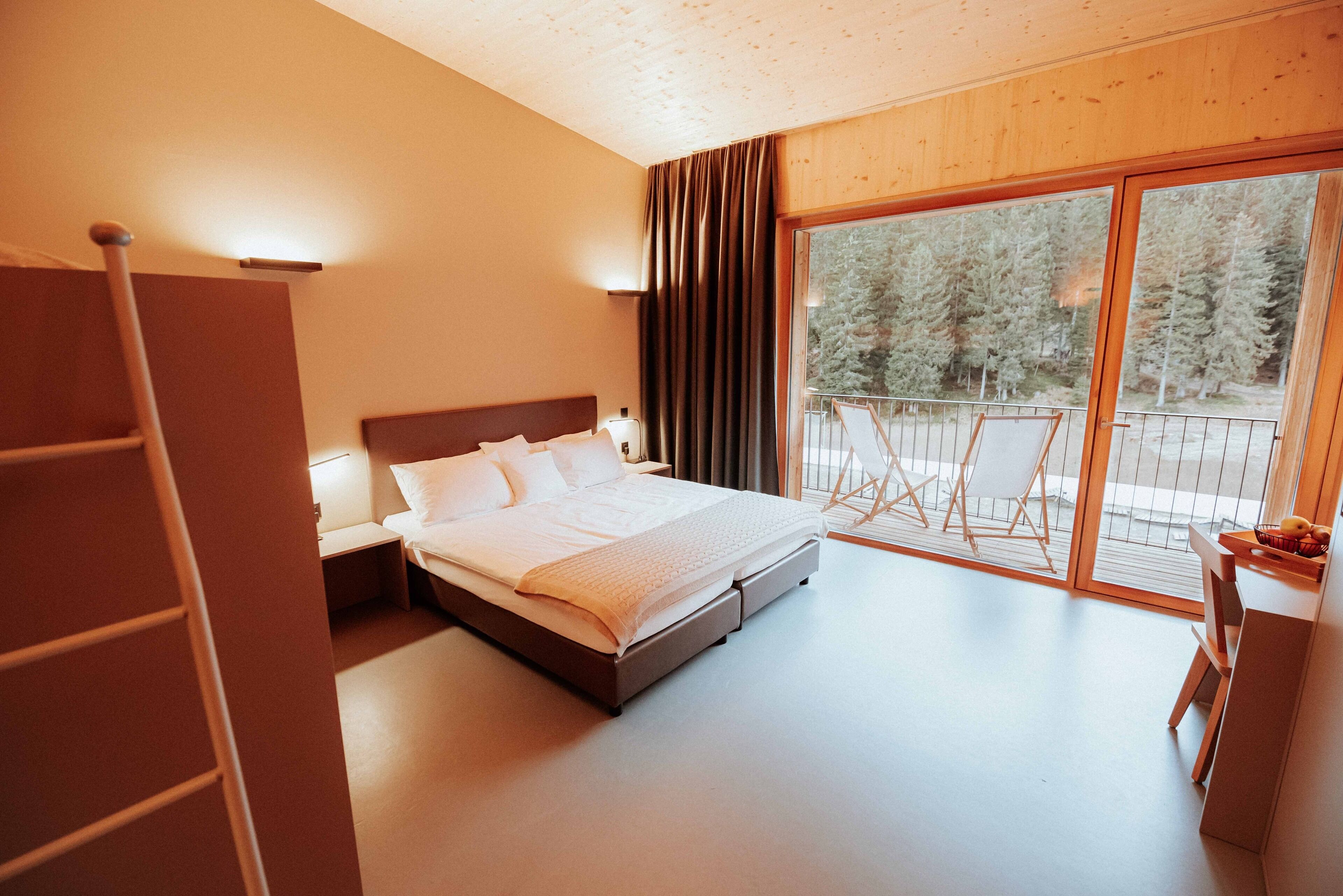 A bedroom with a large bed, a nightstand, a lamp, a wooden wardrobe, and a sliding glass door leading to a balcony with a view of trees.
