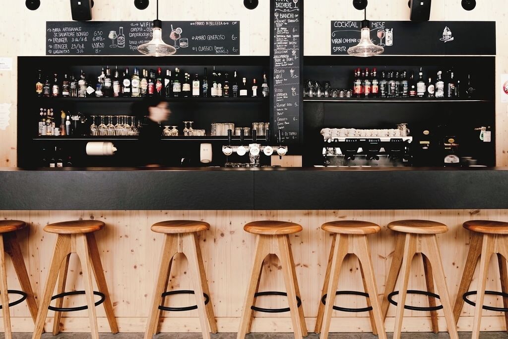 Zona de bar con taburetes de madera, una encimera negra y una pizarra con la carta de bebidas y precios.