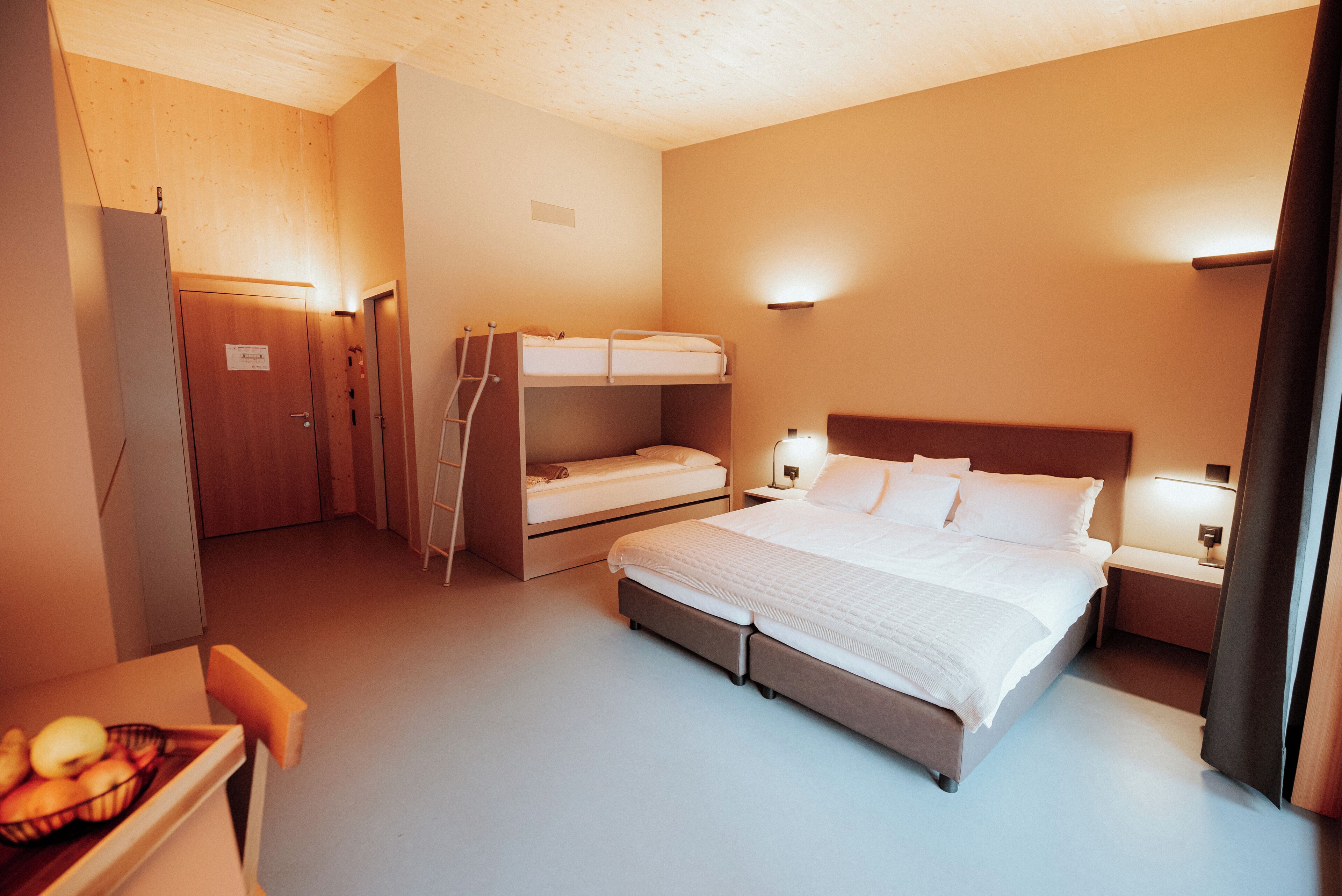 A hotel room with a large bed, a smaller bed on the left, a desk with a chair, and a fruit bowl on the desk.
