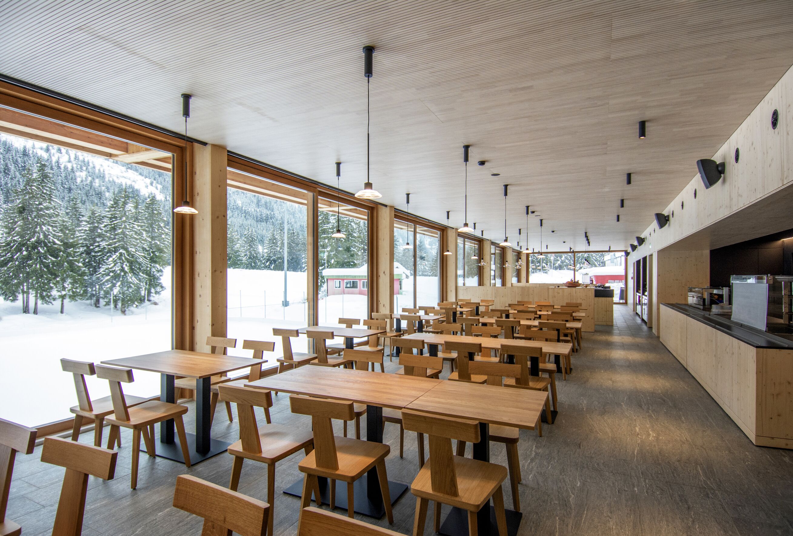 A spacious indoor area with multiple wooden tables and chairs, large windows offering a view of snow-covered trees, and a modern ceiling design.