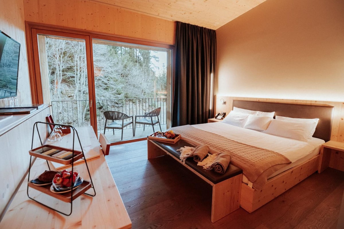 Habitación de hotel con cama, escritorio con silla, una pequeña mesa con comida y vistas a árboles desde la ventana.