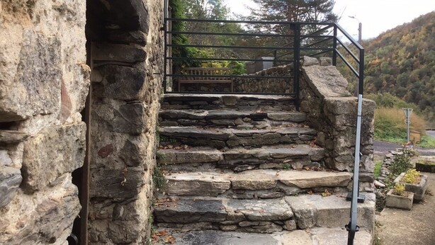 Eine Steintreppe führt zu einem Metalltor, umgeben von Natur und einer Steinmauer.