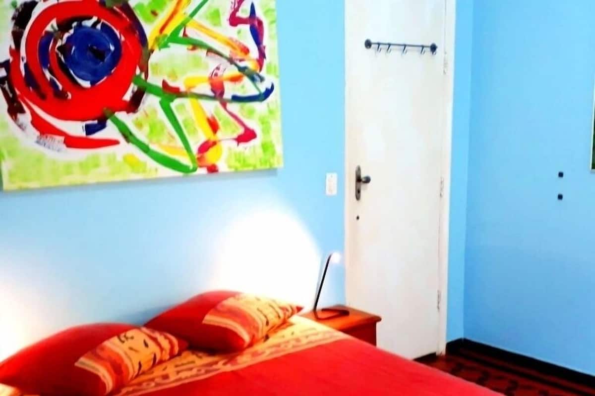 A room with a red bed, a green towel, and a colorful abstract painting on the wall.