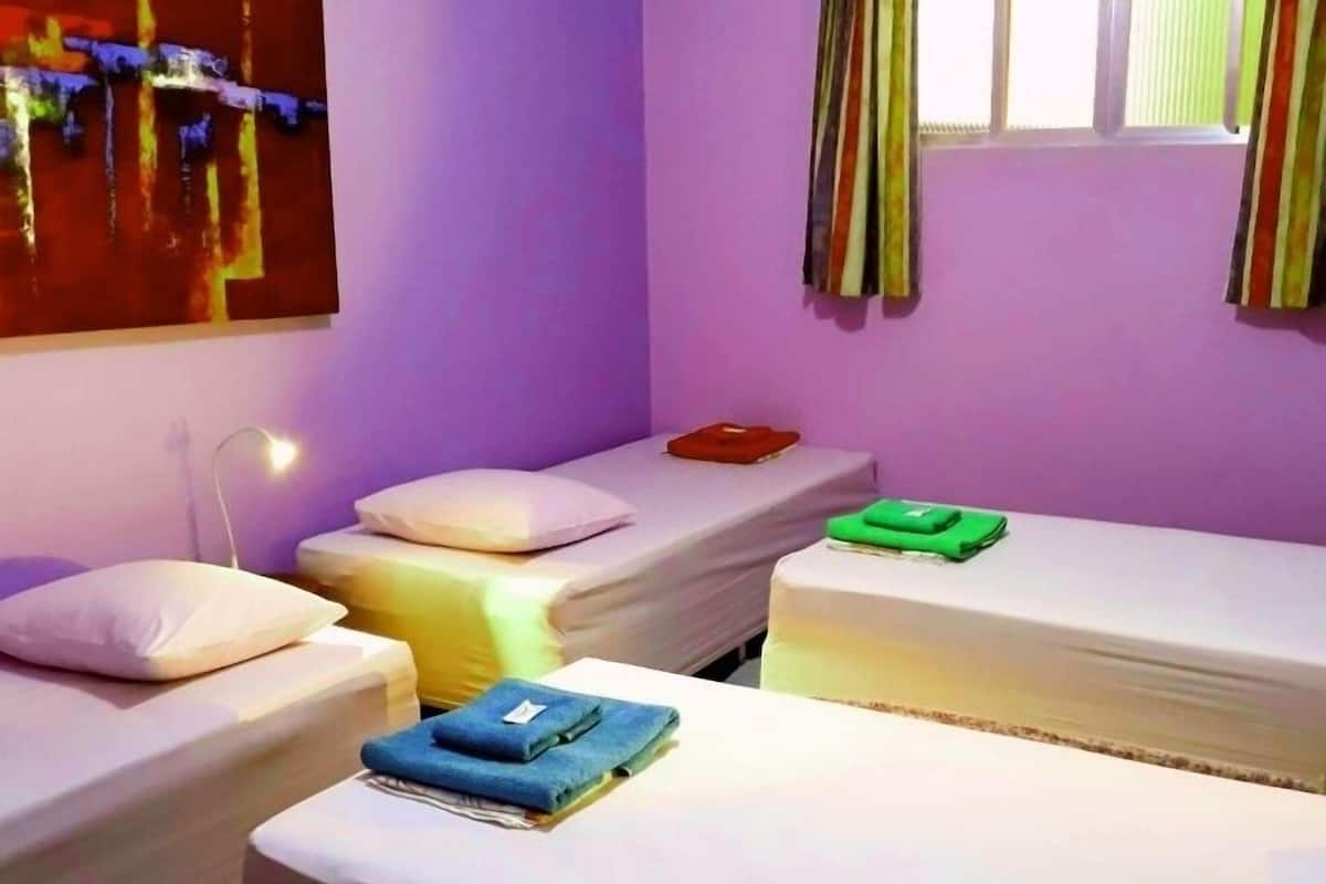 A room with three beds, each with a pillow and a blanket. There is a painting on the wall and a window with curtains.