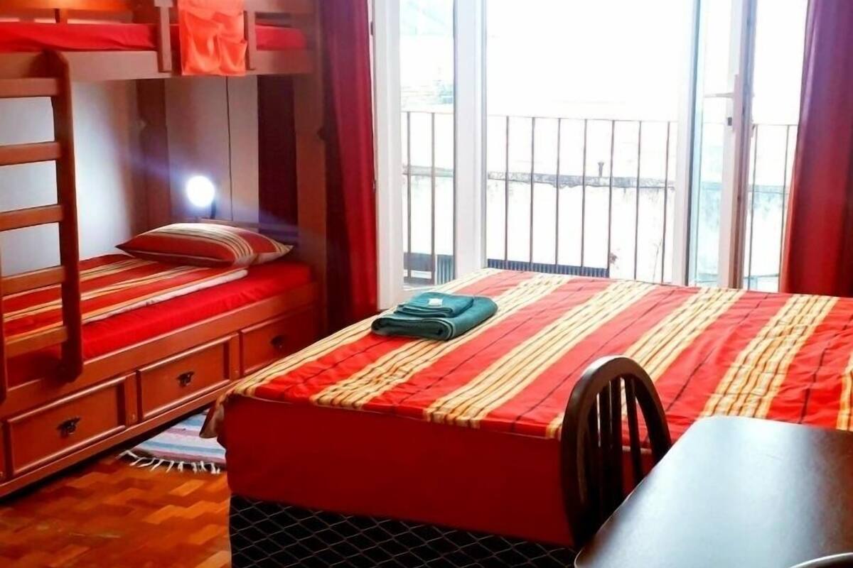 A room with a bunk bed, a single bed, a table, and a chair. There is a window with curtains and a view of the outside.