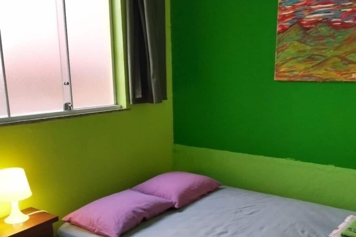 A small, single-bed room with a green wall, a window with curtains, a bedside lamp, and a painting.