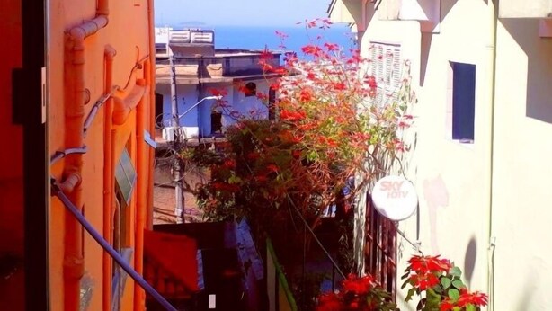 A narrow street with buildings on either side, a clear sky, and a view of the sea in the distance. - Casa Babilônia (Rio de Janeiro)