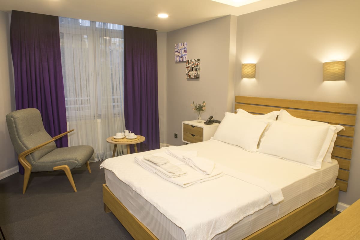A hotel room with a bed, a chair, a small table with a vase, and a nightstand with a phone.