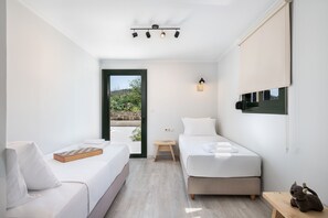A modern bedroom with two beds, a small table, a bench, and a window with a view of greenery.