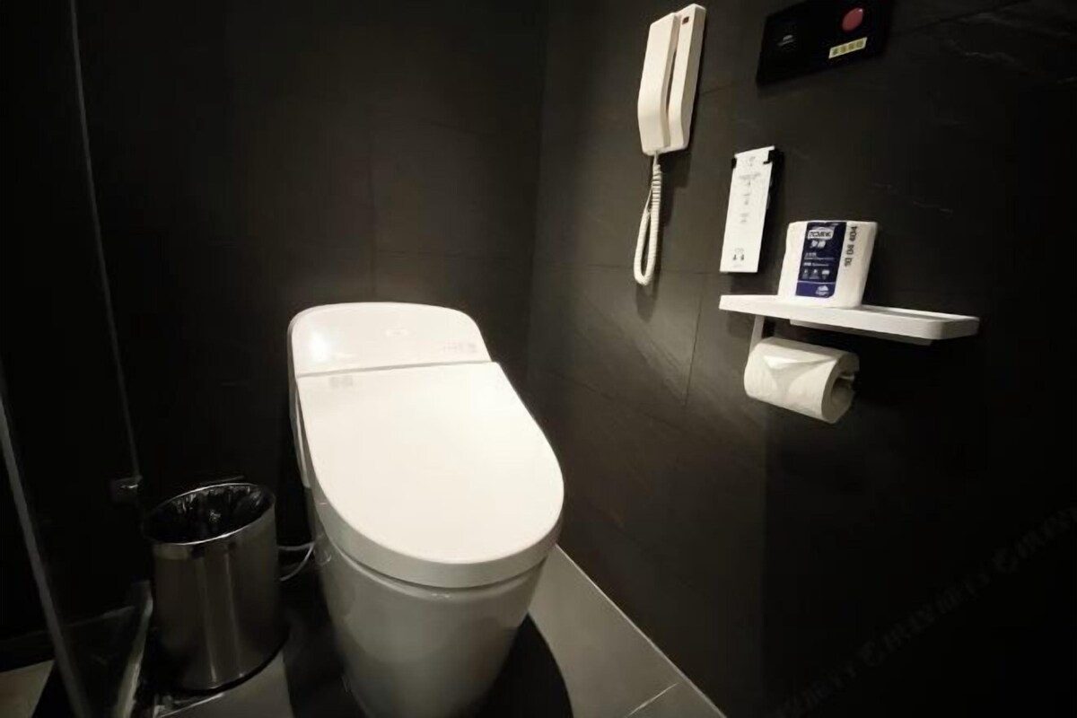 A modern bathroom with a white toilet, a trash bin, a wall-mounted phone, and a toilet paper holder.