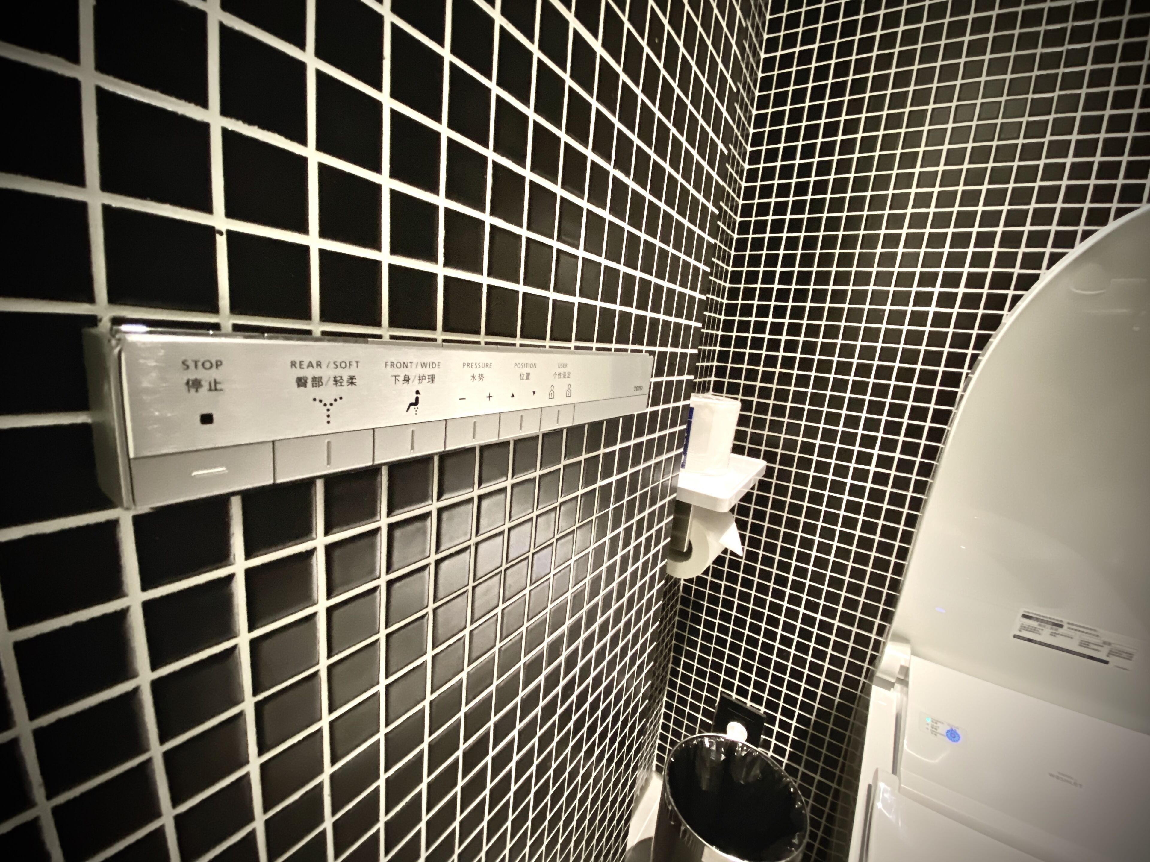 A bathroom with a tiled wall, a hand dryer, and a trash bin.